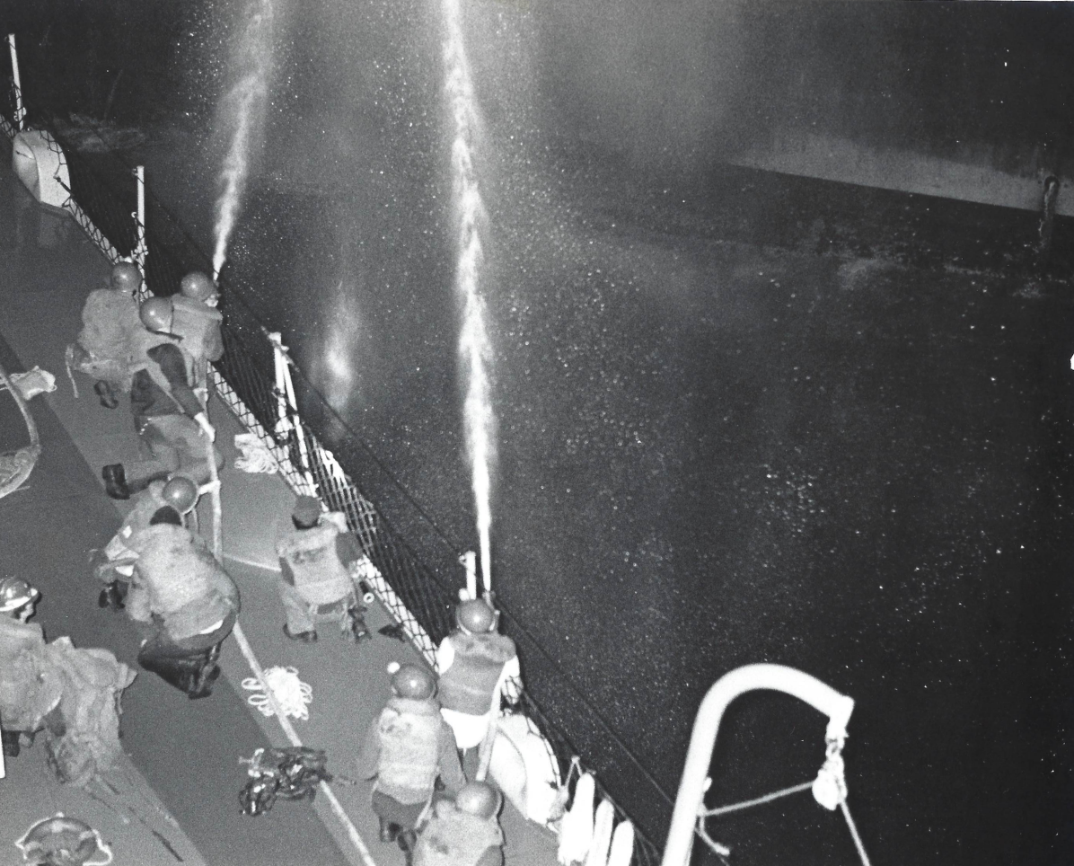 Sailors on the deck of a rescuing ship at night, playing firehoses across the dark water toward USS Belknap.