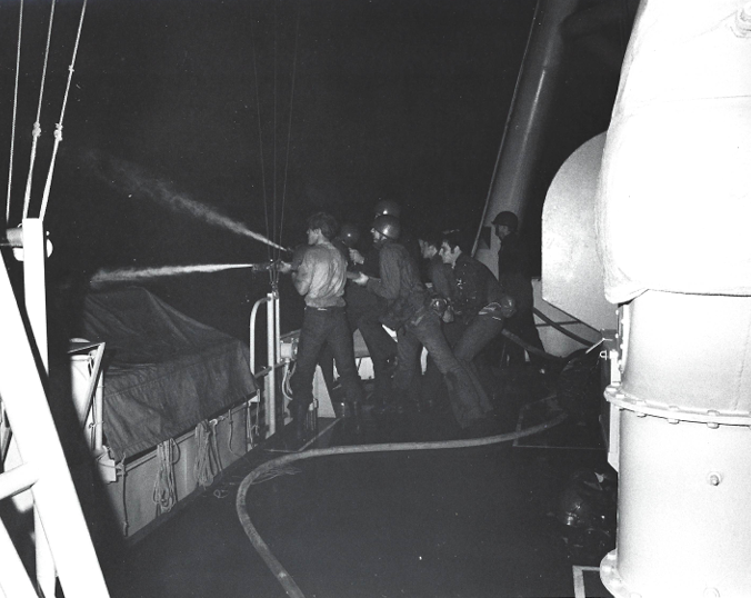 A hose team in helmets and life jackets fighting fire on deck at night, hose lines snaking across the wet steel.